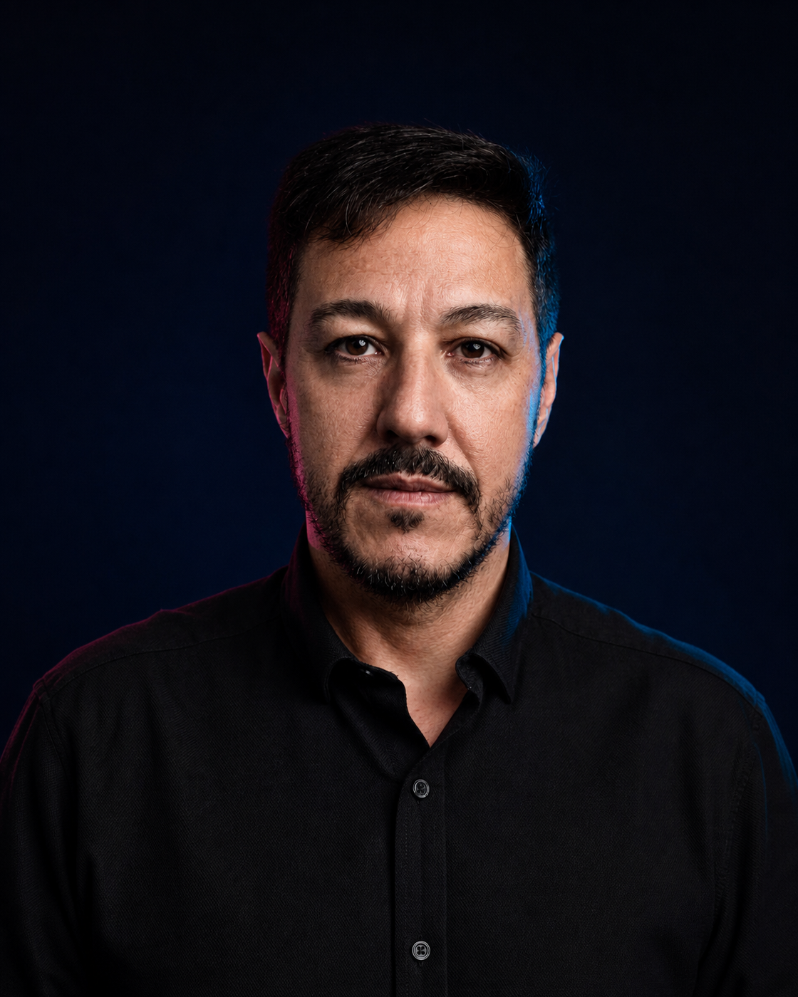 Retrato editorial de Reginaldo Melo, sem óculos, com cabelo curto e barba aparada, sobre fundo navy.